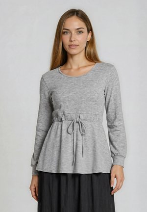 Kobieta ubrana w jasnoszary top z długim rękawem i paskiem w talii oraz czarną plisowaną spódnicę, stojąca na jednolitym tle.