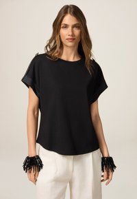 Top negro de manga corta con corte holgado, con un escote redondeado y herrajes decorativos en el hombro. Pulsera de cuentas negras en la muñeca.