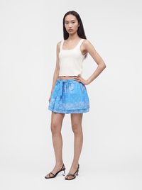 Femme debout, main sur la hanche, portant un top court blanc sans manches, une mini-jupe bleue à motifs avec taille nouée, et des sandales à talons noires.