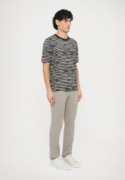 Melns un balts svītrains T-krekls ar apaļu kakla izgriezumu, kopā ar gaiši pelēkām šaurām biksēm un baltām sporta kurpēm, stāvot pret vienkāršu fonu.