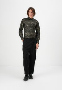 Mann steht mit Händen in den Taschen, trägt ein schwarz-gold gestreiftes Langarmshirt, schwarze weit geschnittene Hosen und schwarze Schuhe vor einem weißen Hintergrund.