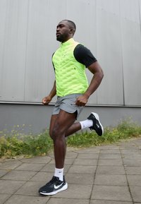 Gilet senza maniche verde neon sopra una maglietta nera, pantaloni corti grigi e scarpe da corsa nere; tessuto testurizzato; uomo che corre su un sentiero pavimentato.