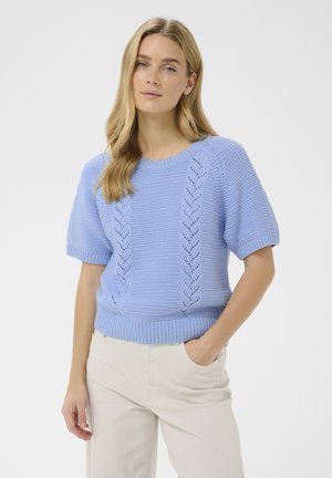 Jeune femme aux cheveux longs et blonds portant un pull tricoté à manches courtes bleu clair et un pantalon beige clair, main dans la poche.