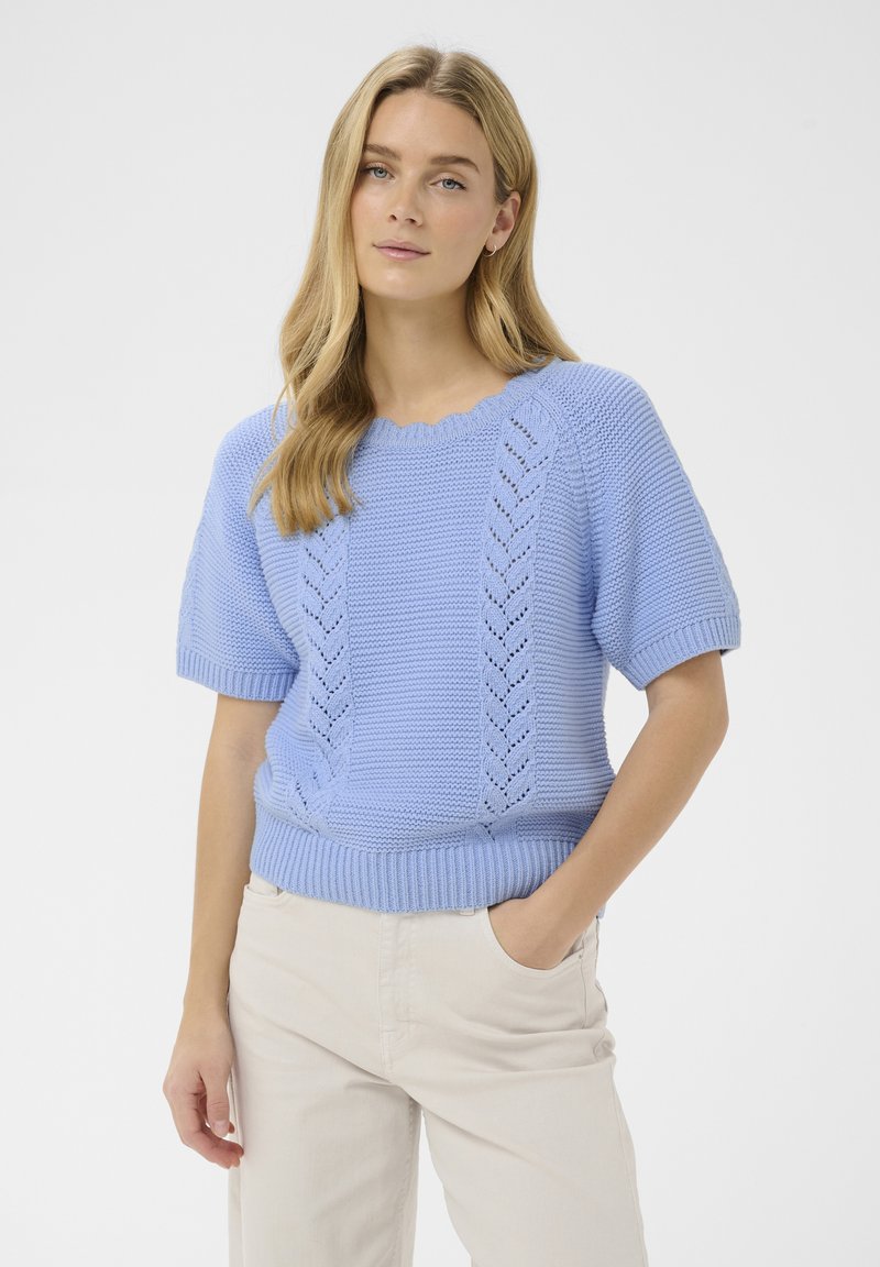 Jeune femme aux cheveux longs et blonds portant un pull tricoté à manches courtes bleu clair et un pantalon beige clair, main dans la poche.