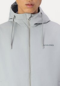 Lichtgrijze hooded jack met een volledige rits, verstelbare trekkoorden en een bedrukte logo op de borst. Glad, textuur stof.