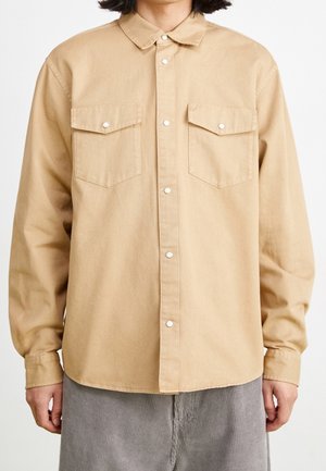 Beige langärmliges Hemd mit Knöpfen und zwei Brusttaschen, getragen von einer Person, kombiniert mit grauer Cordhose.