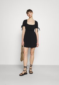 Vestido curto preto com decote quadrado, mangas bufantes e laços. Tecido leve e corte descontraído. Combinado com sandálias pretas de tiras.