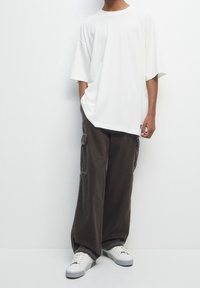 Vit oversized t-shirt, lösa mörkbruna cargo-byxor med sidofickor och vita sneakers med grå detaljer, står mot en neutral bakgrund.