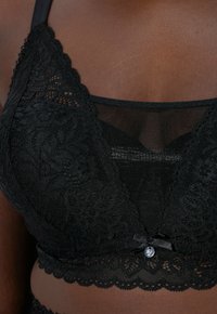 Svart spets-bh med vågiga kanter, blommönster och en genomskinlig meshpanel. Har en liten satinrosett och metallicdetalj.