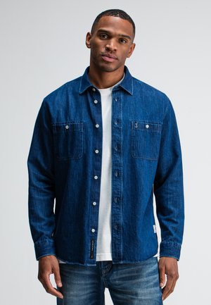 Dunkelblaues Denimhemd mit zwei Brusttaschen, Knopfverschluss und aufgerollten Ärmeln, getragen über einem weißen T-Shirt, vor einem einfarbigen Hintergrund präsentiert.