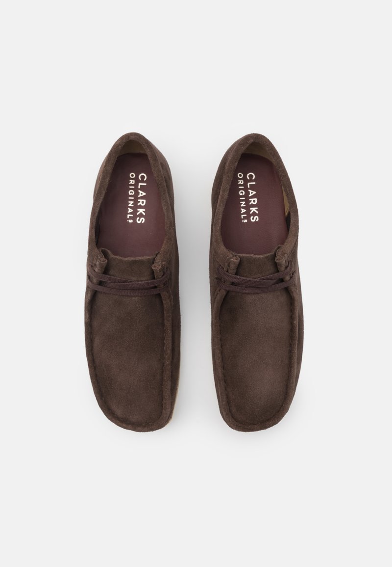 Zapatos slip-on de ante marrón con una textura suave, punta redondeada y detalle de cordones decorativos. Presentan una plantilla con logo.