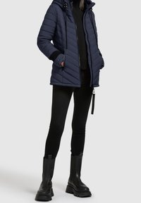 Granatowa pikowana kurtka z kapturem, zamkami błyskawicznymi i kieszeniami. Połączona z czarnymi dopasowanymi spodniami i masywnymi czarnymi butami. Gładka faktura i nowoczesny design.