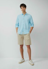 Hellblaues Hemd mit aufgekrempelten Ärmeln, beiger Shorts und weißen Sneakers mit beige Akzenten. Einfaches Design, legere Passform.