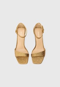 Par de sandalias doradas brillantes con punta abierta y correas para el tobillo, vistas desde arriba sobre un fondo blanco.