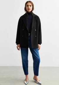Cappotto doppiopetto nero con colletto a vista, indossato sopra un dolcevita blu navy, abbinato a jeans blu dritti e décolleté argentati a punta.