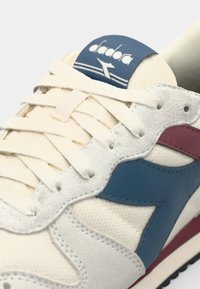 Cream-colored sneaker featuring blue and maroon accents, textured suede and mesh materials, with a cushioned tongue and laces.