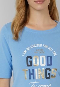 T-shirt en coton bleu clair à manches courtes, avec un motif imprimé en texte blanc, bleu et doré, et un col rond.