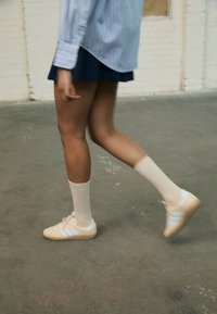 Beige Adidas sneakers med lyseblå striber, båret med hvide knæstrømper. Baggrunden har en betonbund og hvide murstensvægge.
