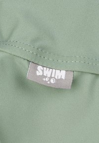 Hellgrüner Stoff mit glatter Textur, der ein graues Etikett zeigt, auf dem das Wort "SWIM" in weißen Bubble-Buchstaben gedruckt ist.