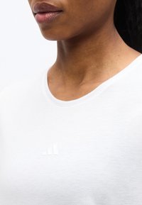 Κοντινό πλάνο του κάτω μέρους του προσώπου και του λαιμού ενός ατόμου που φοράει λευκό πουκάμισο με μικρό, ανάγλυφο λογότυπο Adidas στο στήθος.