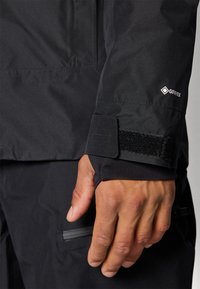 Manica di giacca nera in Gore-Tex con polsino regolabile in Velcro, che mette in mostra una texture liscia e una tasca con zip sul lato.
