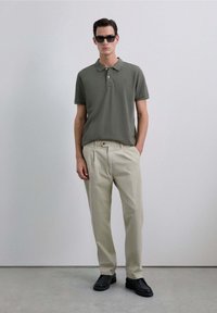 Polo de color verde oliva con mangas cortas y tapeta de tres botones, combinado con pantalones beige claros y zapatos negros sin cordones.