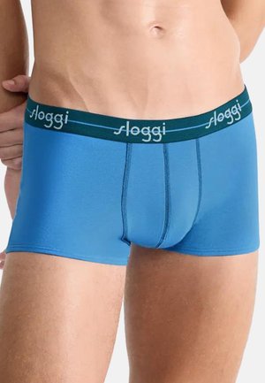 Männlicher Oberkörper, der blaue Sloggi-Boxershorts mit dunkelgrünem Bund und Logo trägt, Hand ruht hinter dem Rücken, weißer Hintergrund.