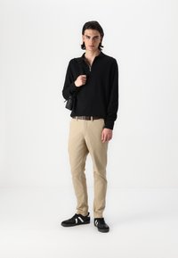Schwarze Zip-Up-Ärmeljacke, beiges Kleid, schwarze Sneakers mit weißen Streifen, hält eine kleine schwarze Tasche und steht vor einem weißen Hintergrund.