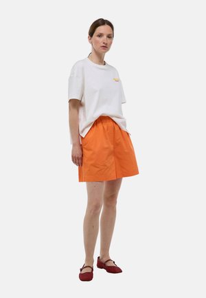 Femme debout portant un t-shirt blanc ample, une jupe orange longueur genou et des chaussures plates rouges, légèrement de profil sur un fond uni.