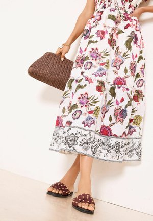 Kobieta w białej sukience midi w kwiatowy wzór, trzymająca teksturowaną brązową kopertówkę i nosząca bordowe klapki z ćwiekami.