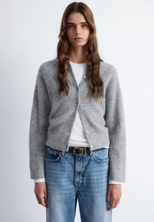 Grauer, knöpfbarer Cardigan aus weichem, strukturiertem Stoff, mit langen Ärmeln und taillierter Passform, über einem weißen Oberteil und blauen Jeans getragen.