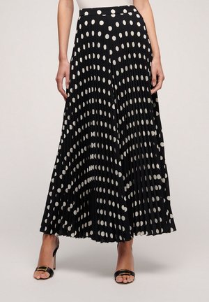 Woman wearing a black pleated midi skirt with white polka dots and black open-toe heels with gold buckles.