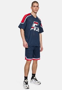 Maglietta sportiva in rete blu navy con accenti rossi, caratterizzata da un audace logo FILA. Abbinata a pantaloni corti navy coordinati e sneakers bianche.