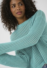 Maglione a maglia di un leggero teal, caratterizzato da un design a trama ampia e aperta, collo a costine e maniche lunghe con una finitura testurizzata.