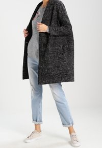 Personne portant un manteau noir texturé, un pull gris avec broderies florales, un jean clair déchiré roulé aux chevilles, et des baskets beiges.