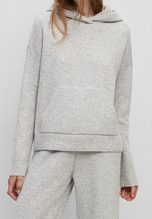 Vrouw draagt een lichtgrijze geribbelde gebreide hoodie en bijpassende wijde broek, staand tegen een effen grijze achtergrond.