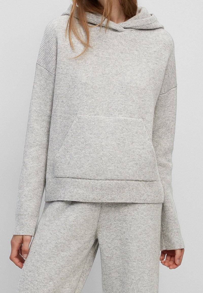 Femme portant un sweat à capuche en maille côtelée gris clair et un pantalon large assorti, debout devant un fond gris uni.