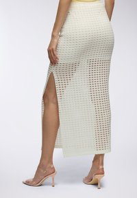 Gonna maxi bianca in rete con uno spacco laterale, caratterizzata da un motivo a griglia e una texture liscia. Abbinata a sandali con tacco trasparenti.