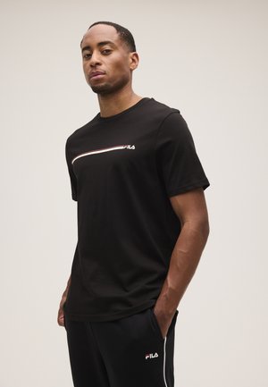 Uomo che indossa una t-shirt e pantaloni neri FILA, in piedi con le mani nelle tasche su uno sfondo chiaro e uniforme.