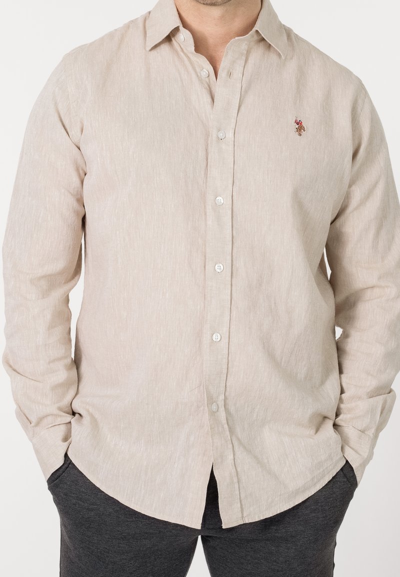 Homme portant une chemise beige clair à manches longues boutonnée avec un petit logo brodé sur la poitrine et un pantalon gris foncé.