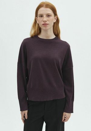 Jeune femme aux cheveux roux portant un pull violet foncé à manches longues et un pantalon noir, debout devant un fond blanc uni.