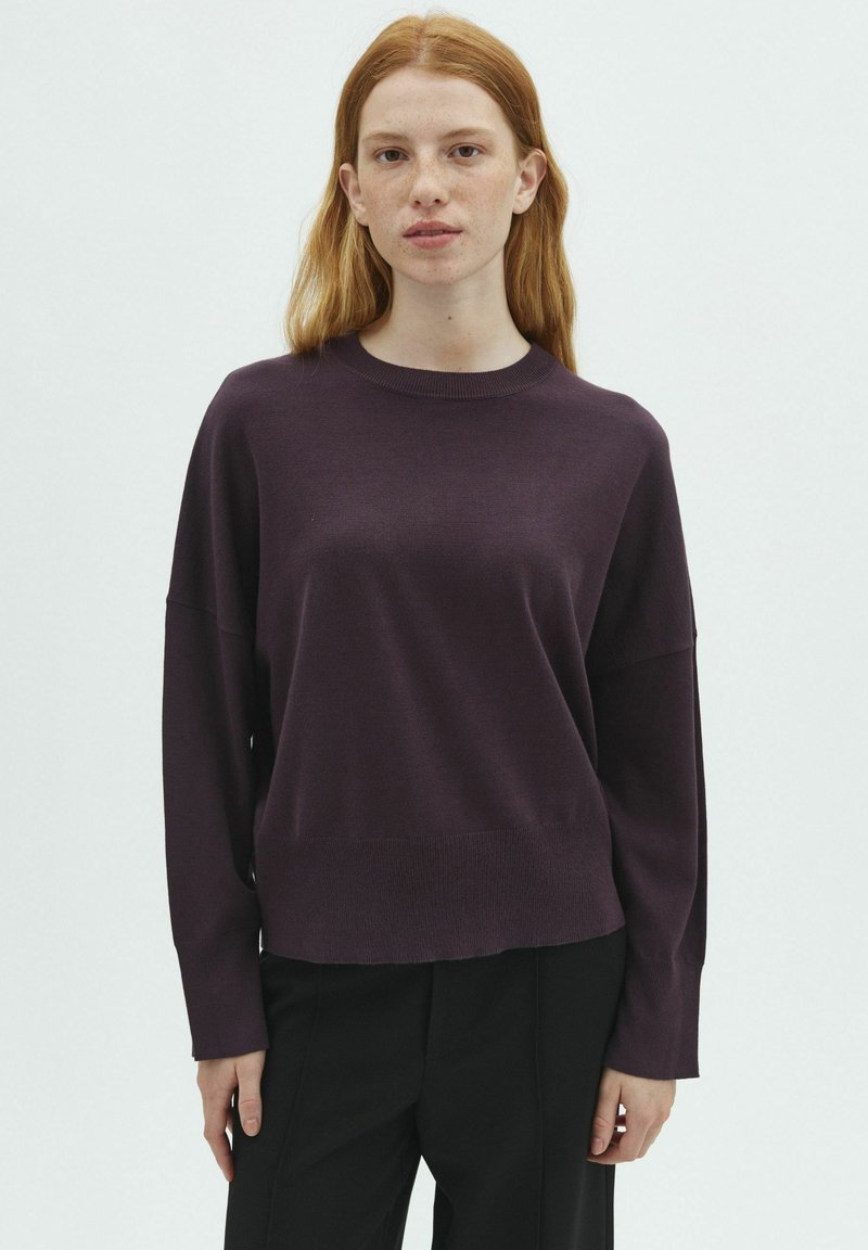 Jeune femme aux cheveux roux portant un pull violet foncé à manches longues et un pantalon noir, debout devant un fond blanc uni.