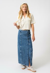 Chemisier blanc raccourci en tissu texturé avec deux poches poitrine, associé à une longue jupe en denim avec des boutons sur le devant et une fente haute.