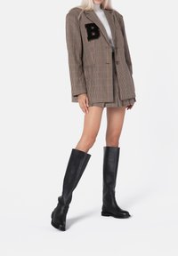 Bruine geruite blazer met zwart "B"-patch, gecombineerd met een bijpassende geruite minirok en zwarte leren laarzen tot de knie. Soepele textuur.