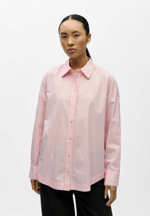 Femme portant une chemise ample rose à boutons avec fines rayures rouges et bords festonnés, associée à un pantalon noir, debout devant un fond blanc.