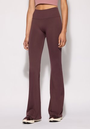 Donna che indossa pantaloni a vita alta e gamba larga di colore malva scuro con sneakers, in piedi davanti a uno sfondo chiaro e uniforme.