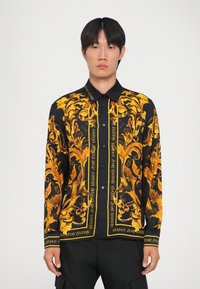 Maglietta nera con ornamenti dorati, dotata di design con bottoni, colletto aperto e testo del marchio a contrasto sul davanti.