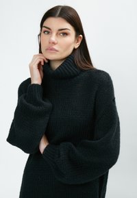 Schwarzer gestrickter Pullover mit hohem Rollkragen, übergroßen Ärmeln und einem strukturierten Muster; das weiche Material sorgt für ein gemütliches Erscheinungsbild.