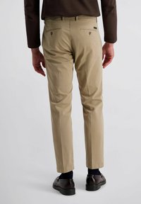 Beige Hose mit schmaler Passform, ausgestattet mit Gesäßtaschen mit Metallknöpfen. Kombiniert mit dunklen Schuhen und marineblauen Socken, von hinten gezeigt.