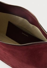 Burgundska kožna torbica sa teksturiranom površinom, zatvaranjem na zatvarač i bež tkaninom unutrašnjosti koja sadrži malu karticu s imenom branda.